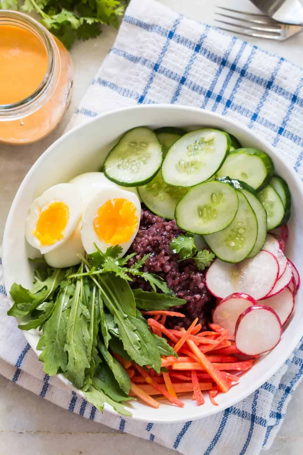 top shot of black rice salad bowl ingredients in a white bowl placed on a blue and white cloth with a jar of dressing on the side