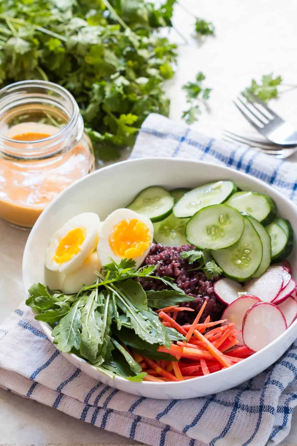 shot of black rice salad bowl ingredients in a white bowl placed on a blue and white cloth with forks, coriander and a jar of dressing on the side