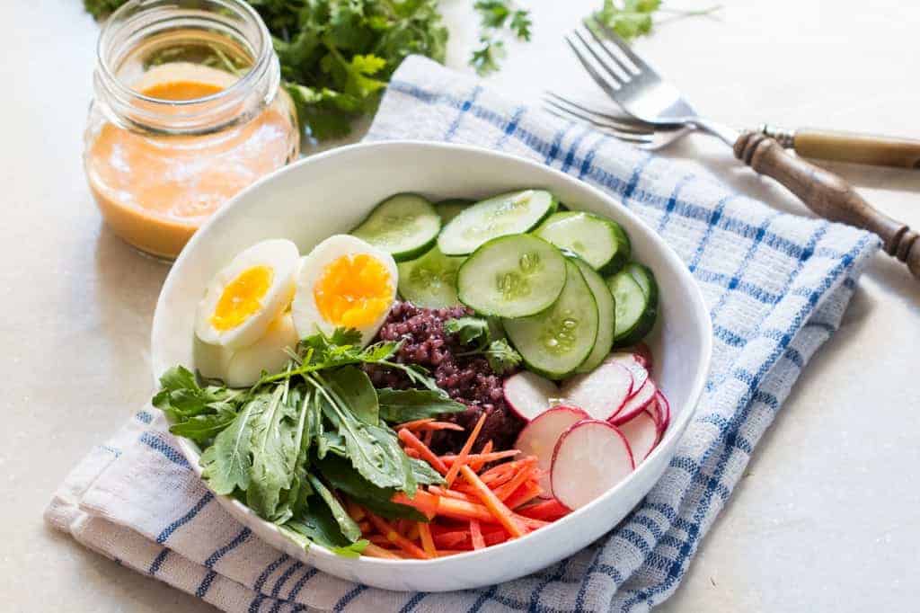 shot of black rice salad bowl ingredients in a white bowl placed on a blue and white cloth with forks, coriander and a jar of dressing on the side