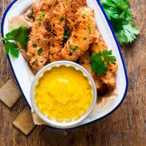 Baked Coconut Chicken Tenders garnished with coriander and served with Mango Mustard Sauce on a white plate.