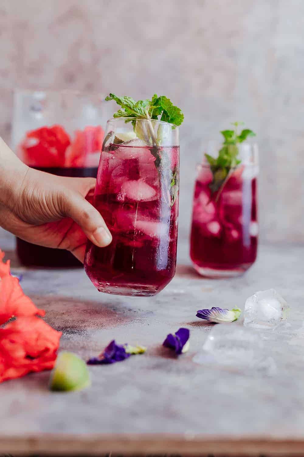 Learn how to make hibiscus tea which has some amazing benefits. Make this with either fresh or dried flowers and choose to have it as a hot cup of tea or as hibiscus iced tea because either way it's delicious and really good for you! #hibiscus #healthfood #drink #icedtea #summer #recipe Picking up a glass of chilled hibiscus tea which is served with mint leaves and lemon