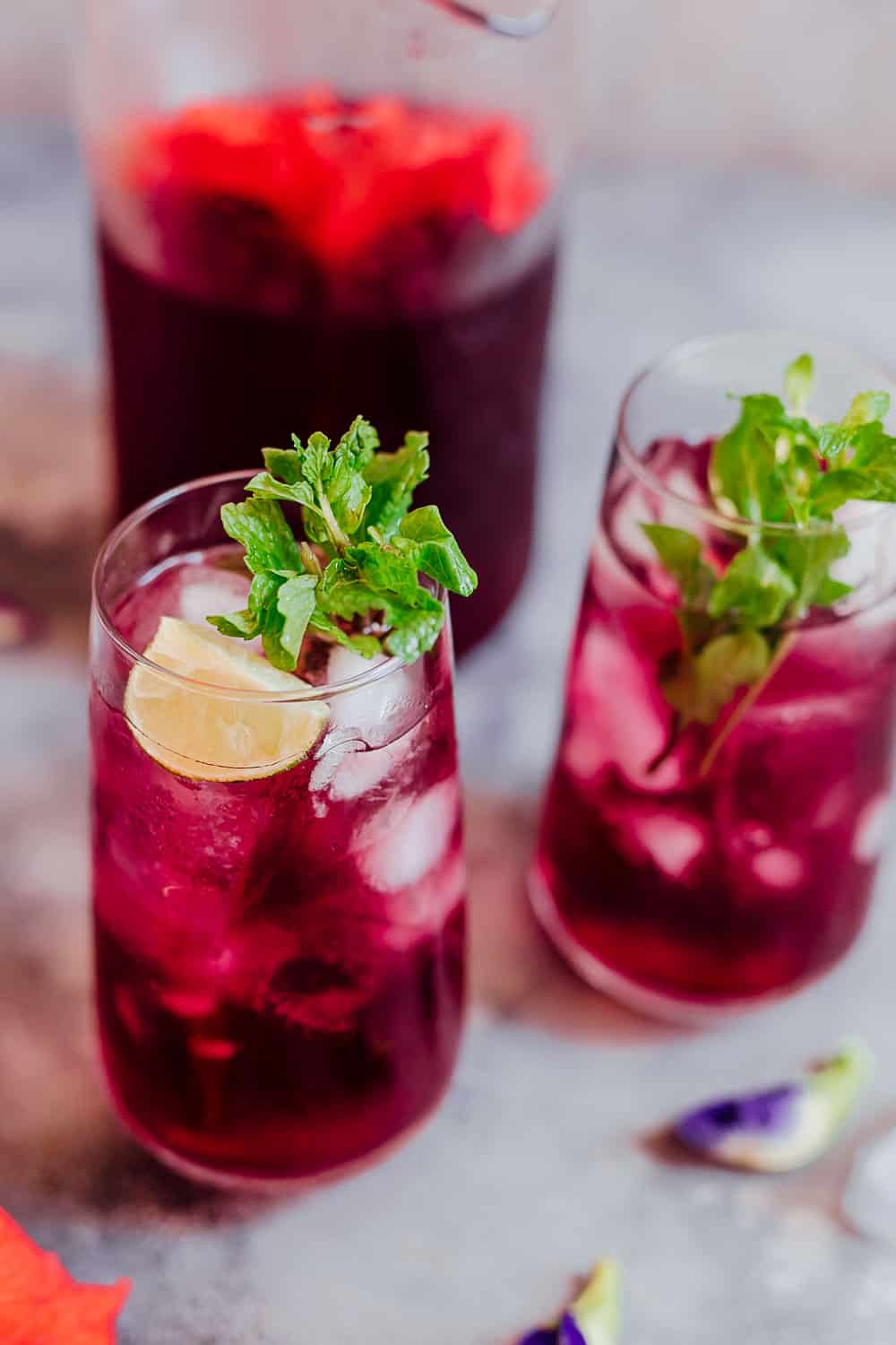 Learn how to make hibiscus tea which has some amazing benefits. Make this with either fresh or dried flowers and choose to have it as a hot cup of tea or as hibiscus iced tea because either way it's delicious and really good for you! #hibiscus #healthfood #drink #icedtea #summer #recipe Closeup of a glass of hibiscus tea with ice cubes, mint and lemon