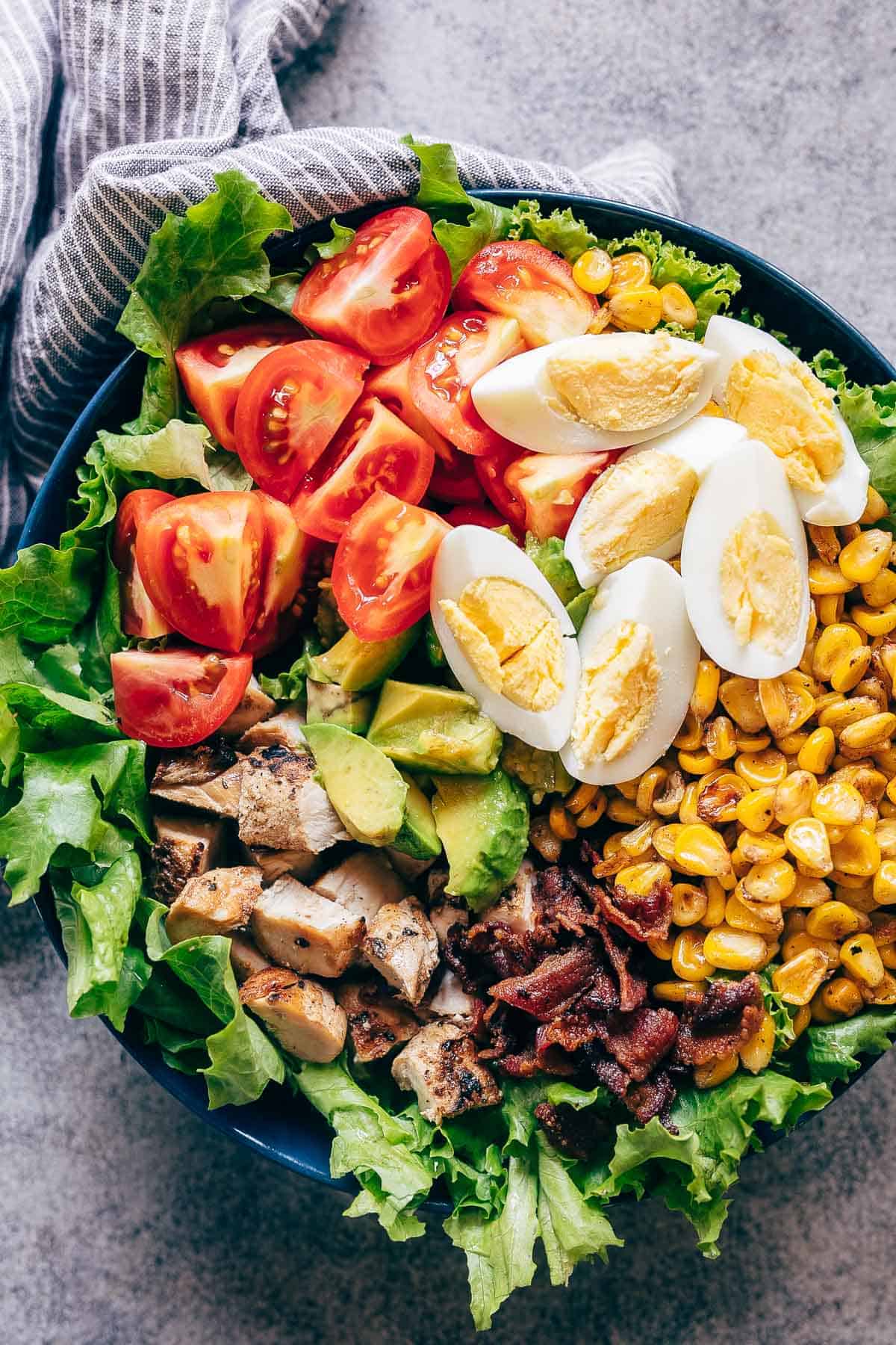 This is a classic layered chicken cobb salad recipe that has all the right fixings that includes lettuce, boiled eggs, grilled corn, bacon, grilled chicken, tomatoes and avocado! Serve this with a creamy avocado blue cheese dressing that's made with greek yogurt and you have dinner sorted. This isn't just a salad, it's a meal! Perfect for a crowd. Use steak instead of chicken or make it vegetarian #salad #recipe #chicken #dinner #easy Chicken Cobb Salad served in a bowl - it has lettuce, tomatoes, chicken, bacon, corn and boiled eggs
