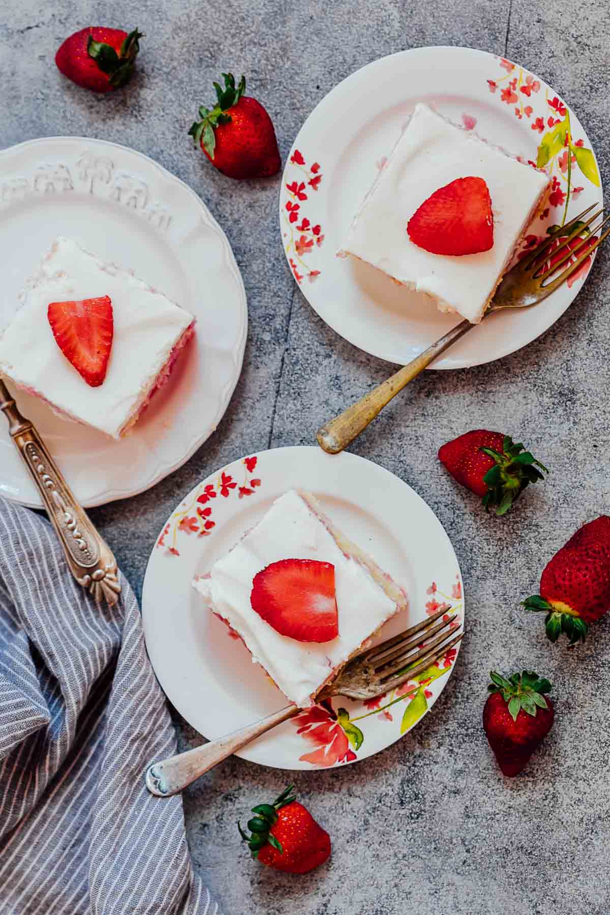 This strawberry poke cake is made from scratch and is a soft, moist jello poke cake that is topped with whipped cream and fresh strawberries. It has the taste of strawberries in every bite and is the perfect easy, elegant dessert! #dessert #cake #strawberry #easter #mothersday #pink Three slices of strawberry poke cake made from scratch served on white plates with forks and fresh strawberries on the side