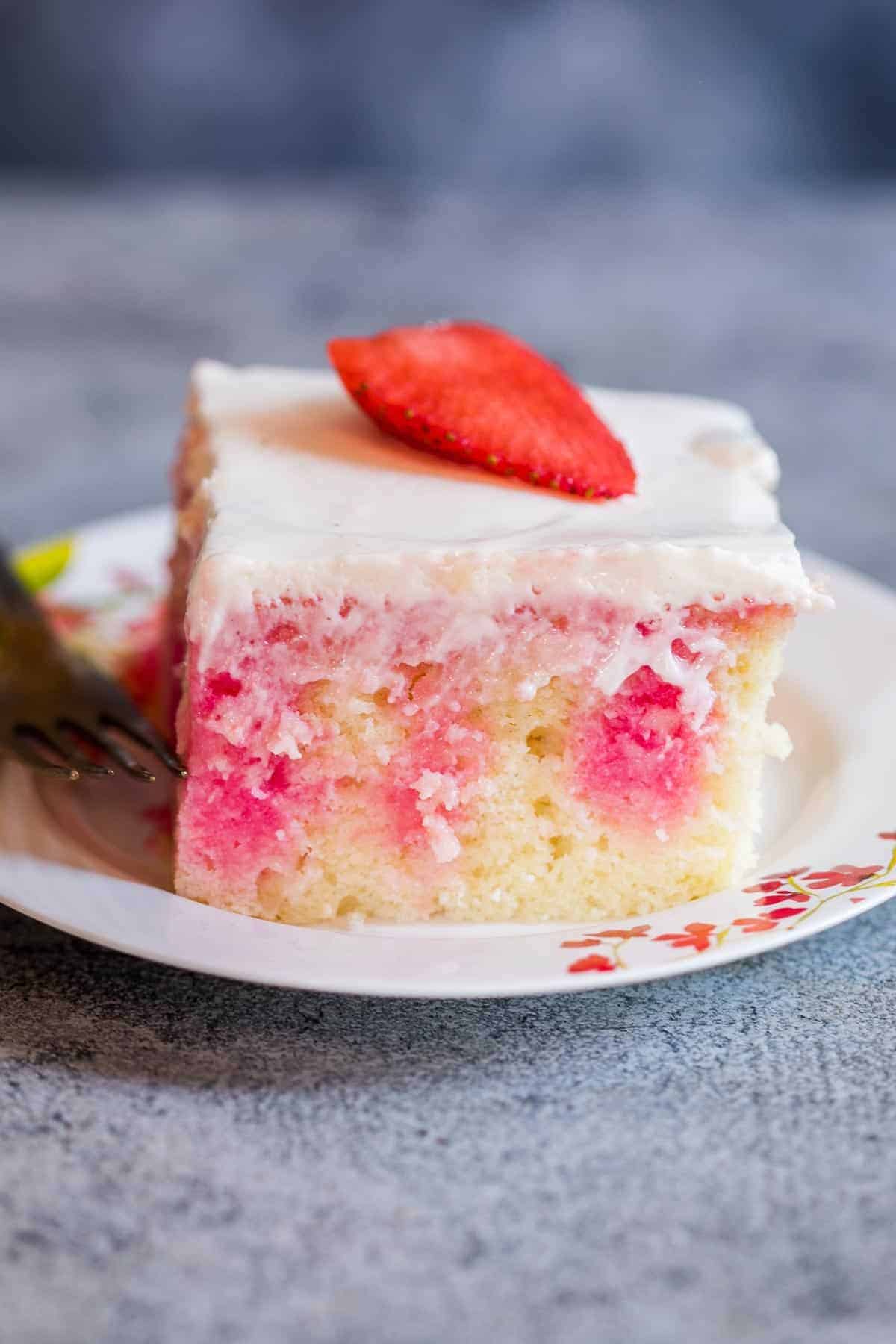 This strawberry poke cake is made from scratch and is a soft, moist jello poke cake that is topped with whipped cream and fresh strawberries. It has the taste of strawberries in every bite and is the perfect easy, elegant dessert! #dessert #cake #strawberry #easter #mothersday #pink Closeup of a slice of a strawberry poke cake with drizzles of jello visible through the cake.