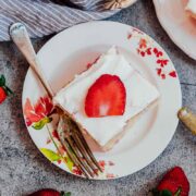 A slice of strawberry poke cake made from scratch served on white plates with forks and fresh strawberries on the side