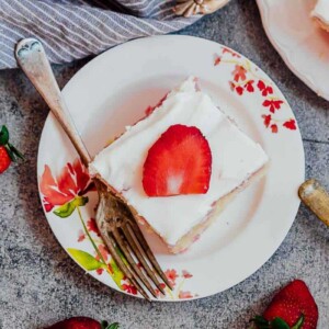 A slice of strawberry poke cake made from scratch served on white plates with forks and fresh strawberries on the side