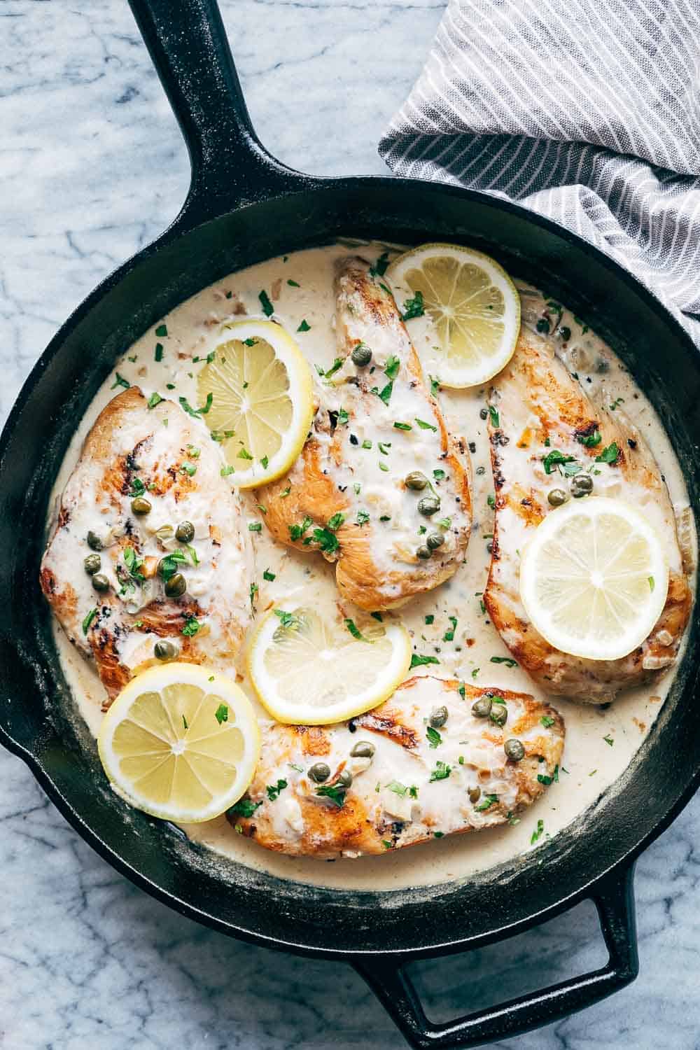 LEMON CHICKEN SCALLOPINI is a super quick, healthy and fast dinner recipe that takes less than 30 minutes to put on the table and has a delicious lemon sauce that you'll want to drink straight from the pan! Studded with capers, this dish looks fantastic. Its gluten free and keto friendly. #fastdinner #chicken #lunch #lemon #summer Creamy lemon chicken scallopini served in a cast iron skillet with four chicken breasts with capers and lemons on top