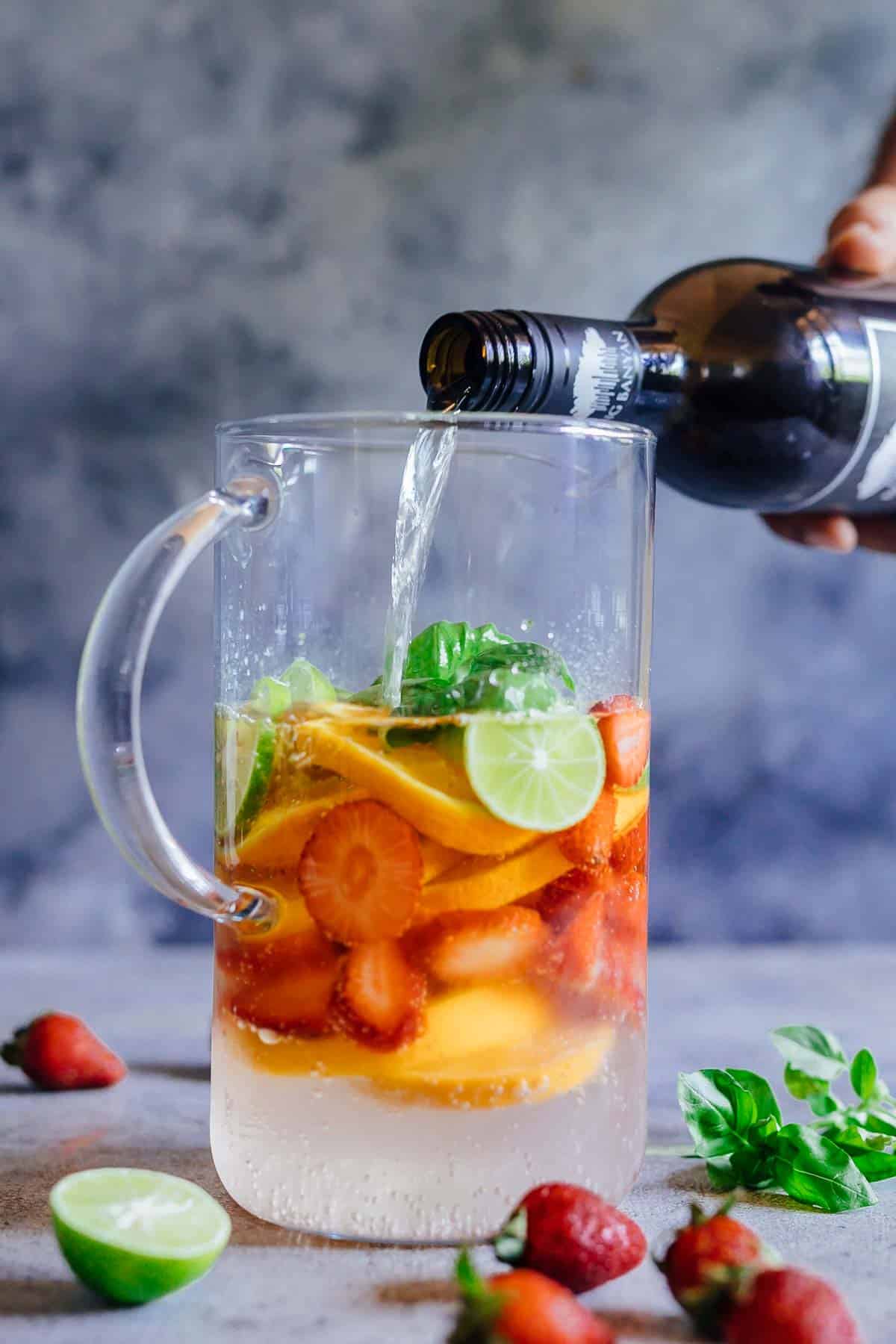 Strawberry Orange White Wine Sangria is a refreshing cocktail that combines the sweetness and flavours of strawberries, oranges, limes and basil with white wine for a fresh take on a traditional sangria. It's the perfect brunch drink that can be made ahead for a crowd. #whitesangria #redwine #cocktails #recipe #drink #summer Pouring wine in a pitcher over fruits to make Strawberry Orange White Wine Sangria