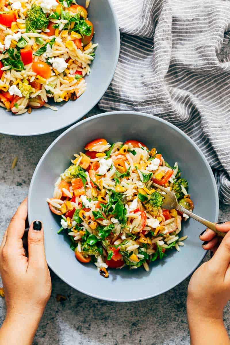 Live your best summer life with this zippy lemon orzo pasta salad! Served cold, its a vegetarian salad that has all the right summer vibes with cherry tomatoes, broccoli, lettuce, corn and bell peppers. Perfect pasta salad for a crowd, and tastes best when made ahead. #salad #pasta #picnicrecipes #potluck #barbecue #cleaneating #lunch Two bowls of colourful, zippy, summery lemon orzo pasta salad with a hand holding one of the bowls