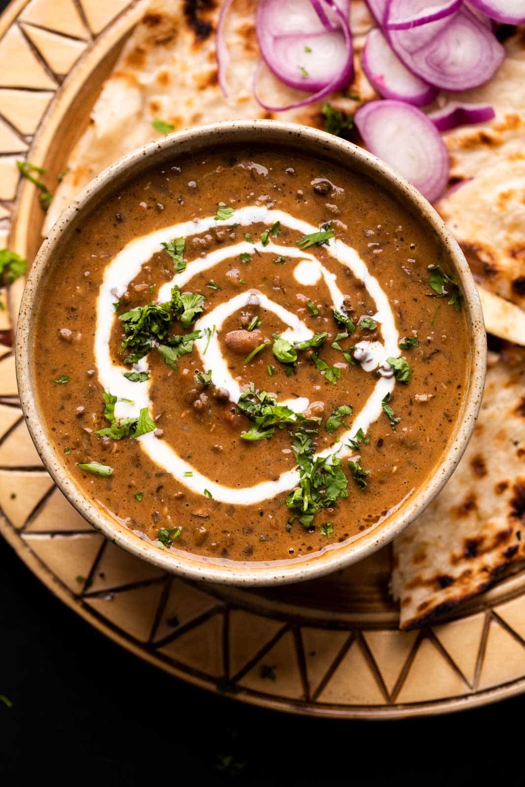 Dal Makhani served in a brown ceramic bowl with cream drizzled on top and naan and sliced onions on the side