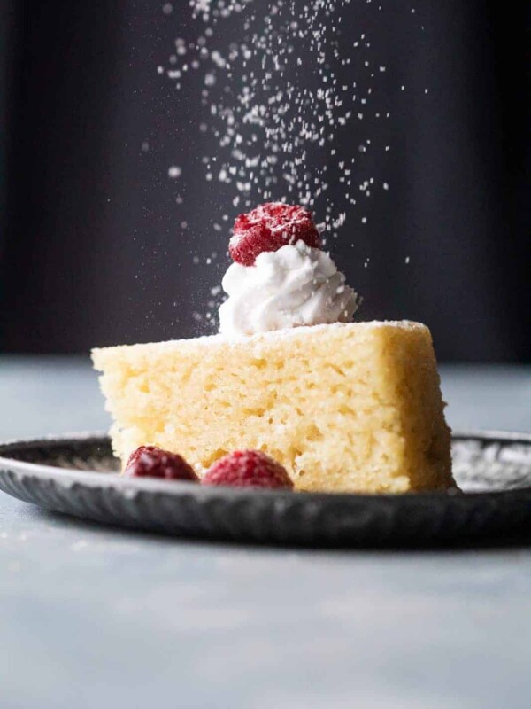 A slice of eggless vanilla cake with whipped cream, raspberries and a sprinkling of icing sugar