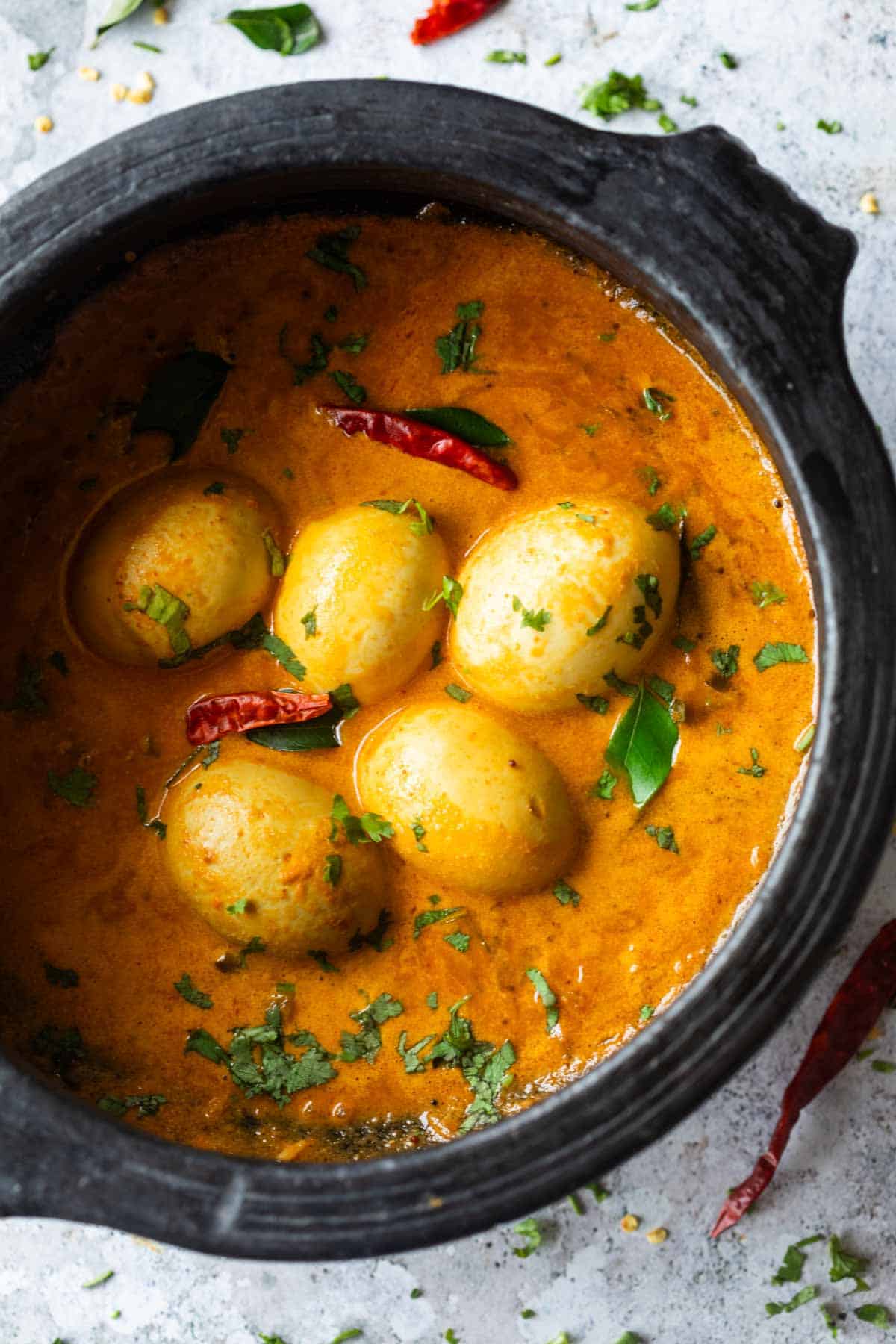 South Indian style Egg Curry is incredibly easy to make. A few spices, onions, tomatoes and coconut milk make this utterly satisfying and comforting egg curry recipe that tastes best with parathas (flatbread) and rice. Make extras because everyone will want seconds! #indian #curry #recipe #coconut #eggs an image of south indian style egg curry in a black clay pot and garnished with coriander and curry leaves