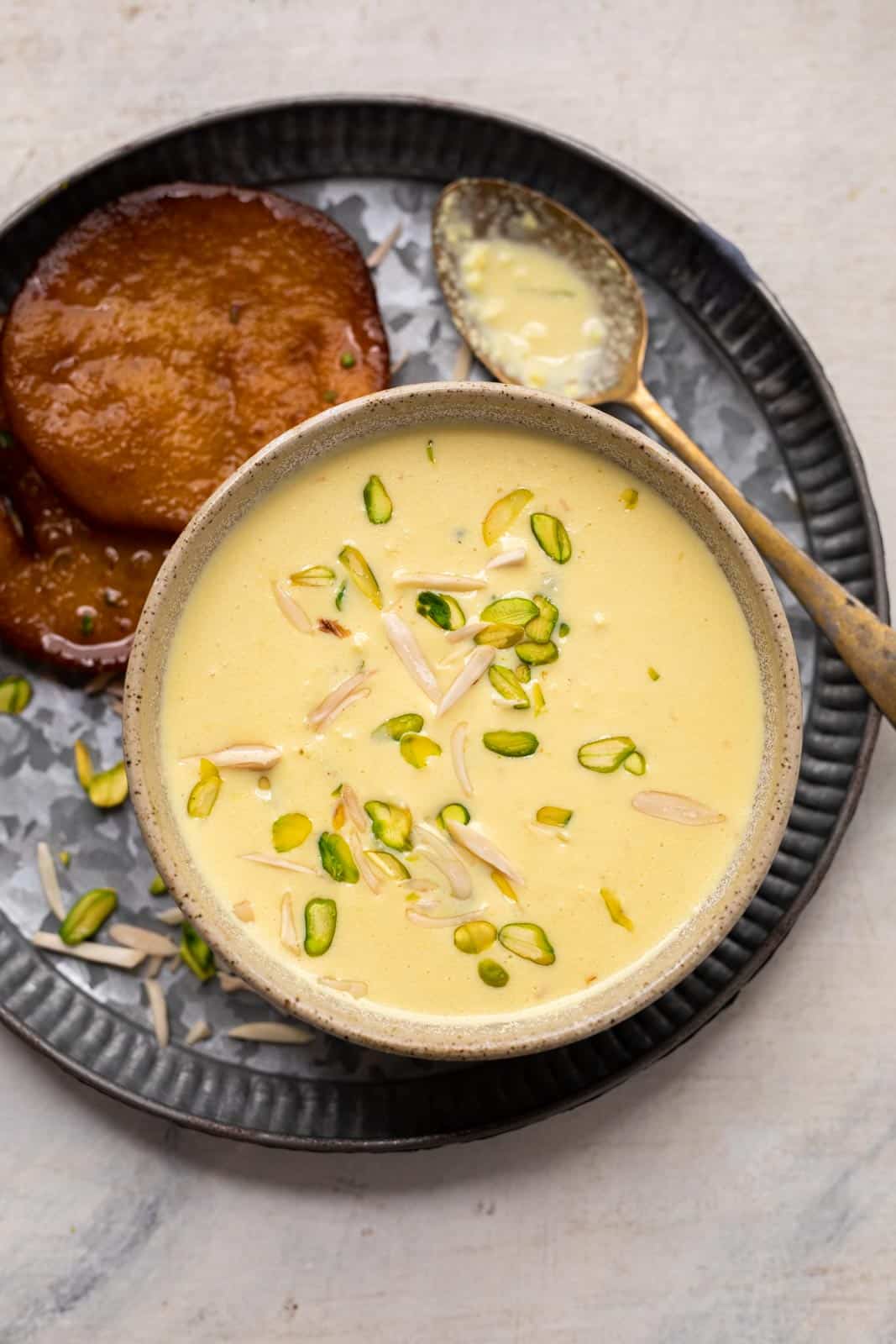 Instant rabdi garnished with slivered almonds and pista served in a brown bowl with malpua on the side