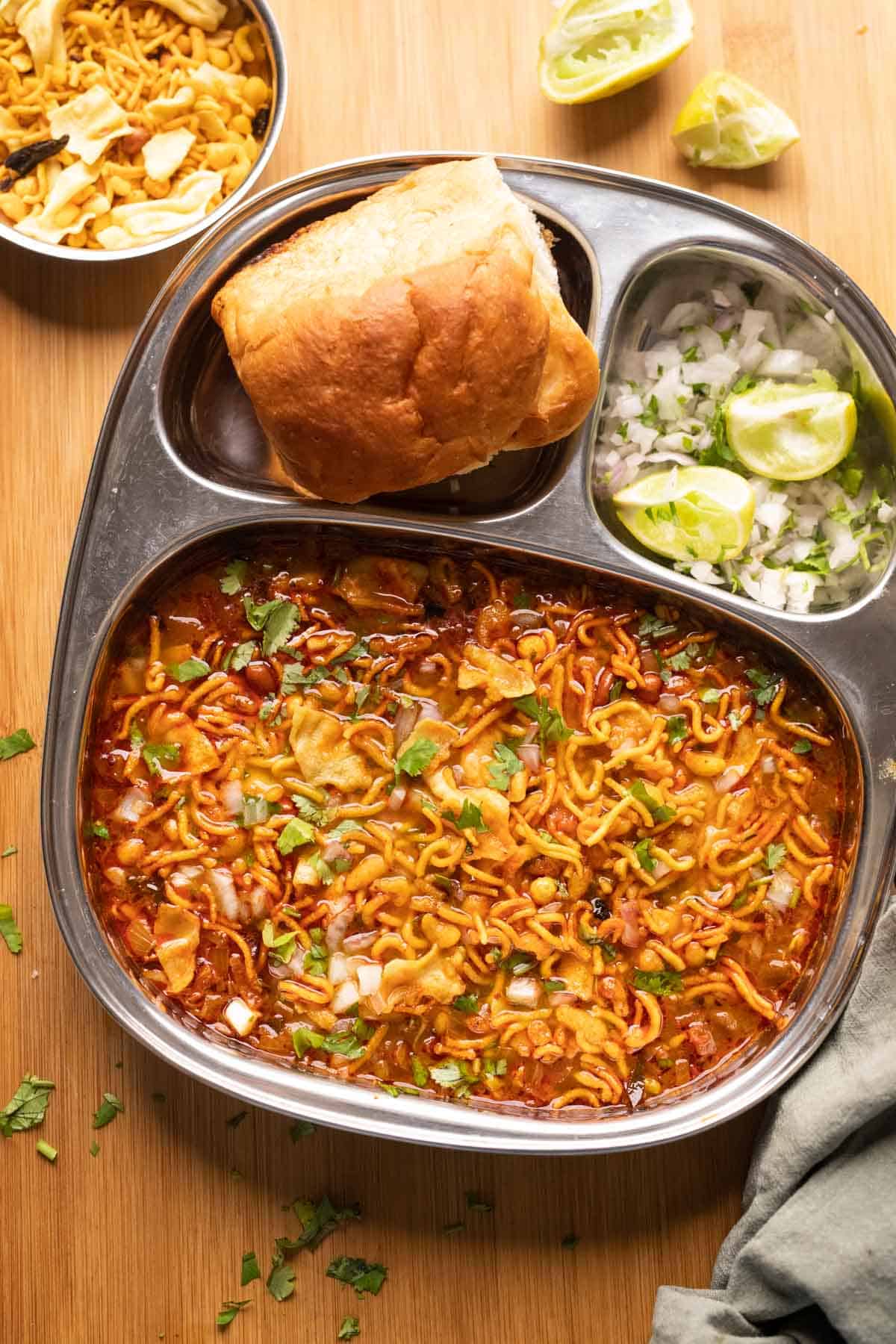 Picture of misal topped with farsaan served in a steel plate with pan, chopped onions and lime wedges on the side