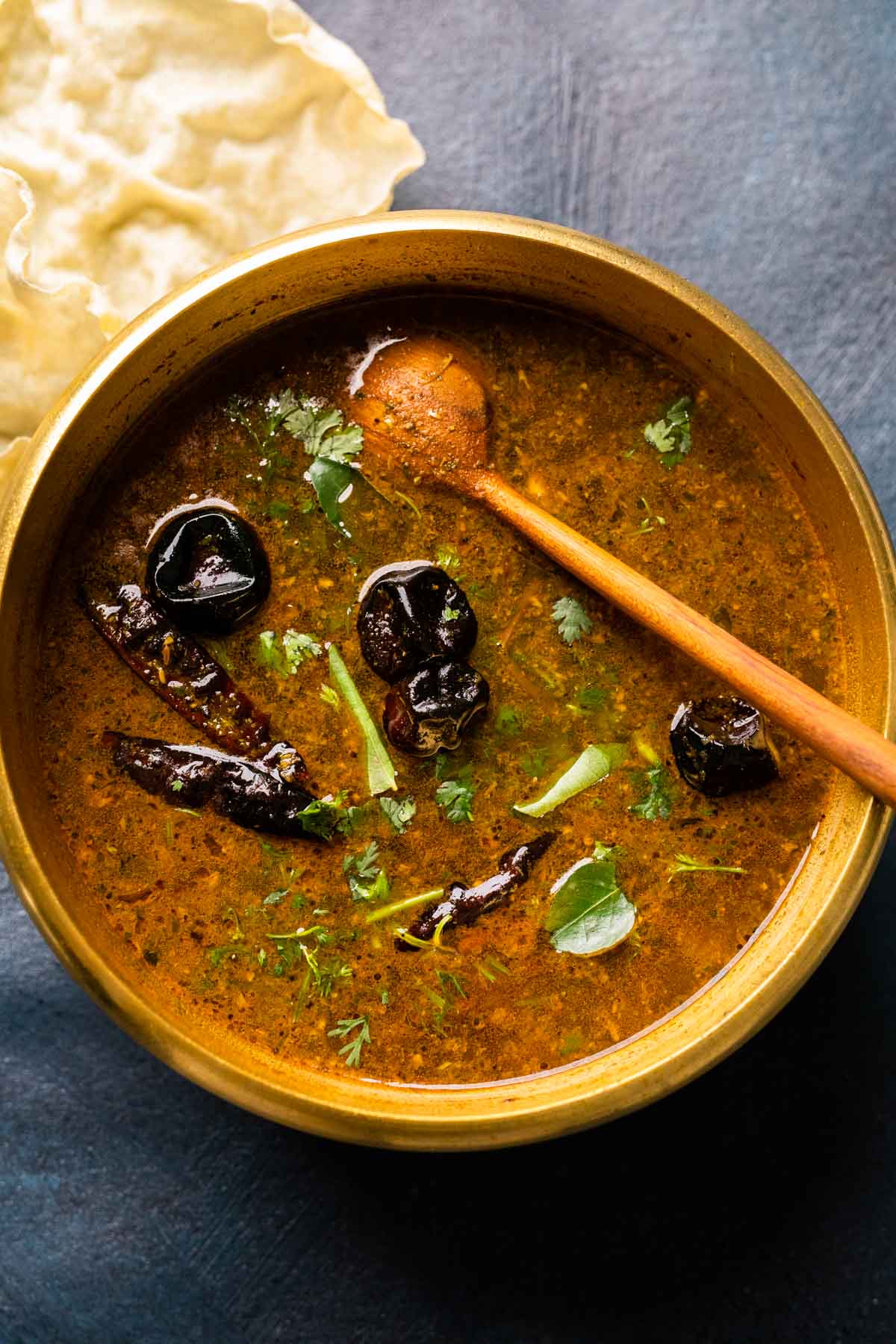 Picture of rasam in the brass pot that it was cooked in with a wooden ladle and some papad on the side