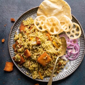 Rice bath served in a grey plate with fryums, poppdum and onions on the side