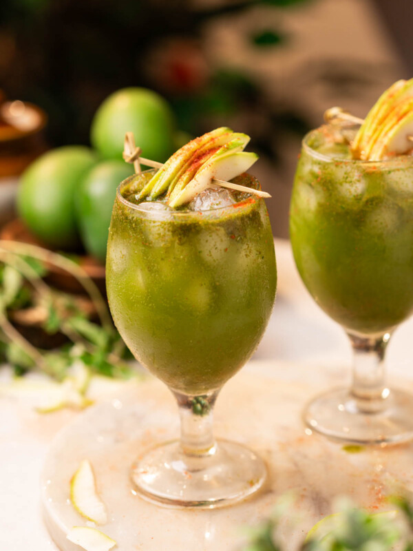 two glasses of aam panna garnished with raw mango slices and chilli powder
