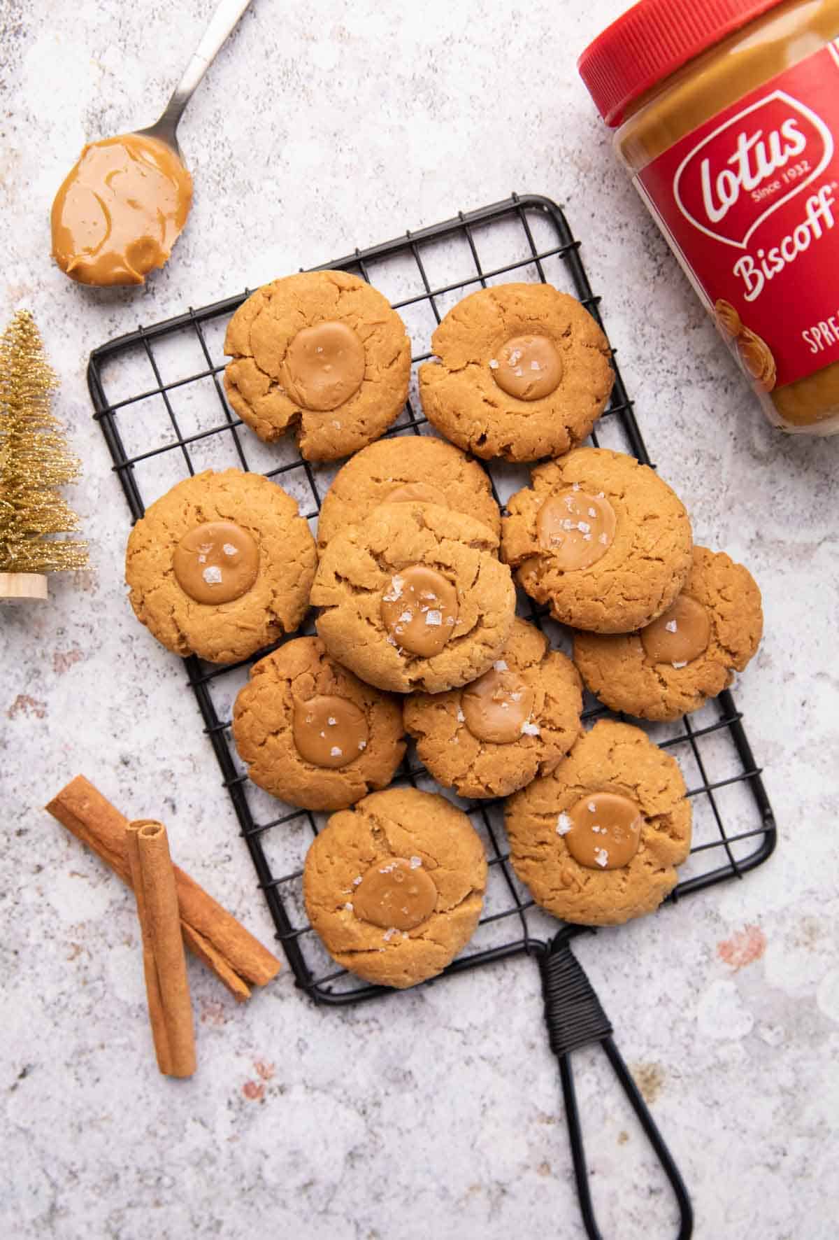 A shot of multiple biscoff thumbprint cookies stacked one on top of another on a black grill along with a spoon of biscoff, a golden christmas tree, two cinnamon sticks and a jar of biscoff on the sides of the grill.