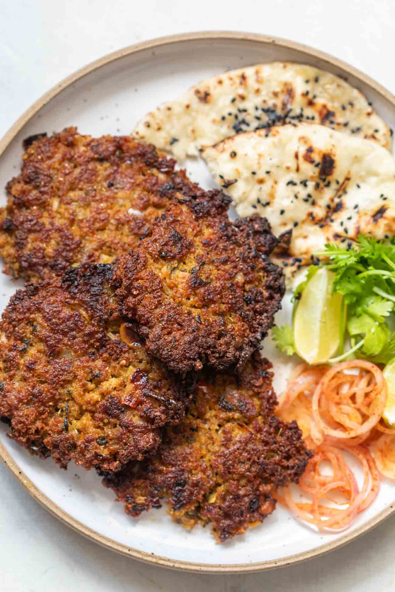 top shot of chapli kebabs served in a white plate with naan, onion, lemon, and coriamder