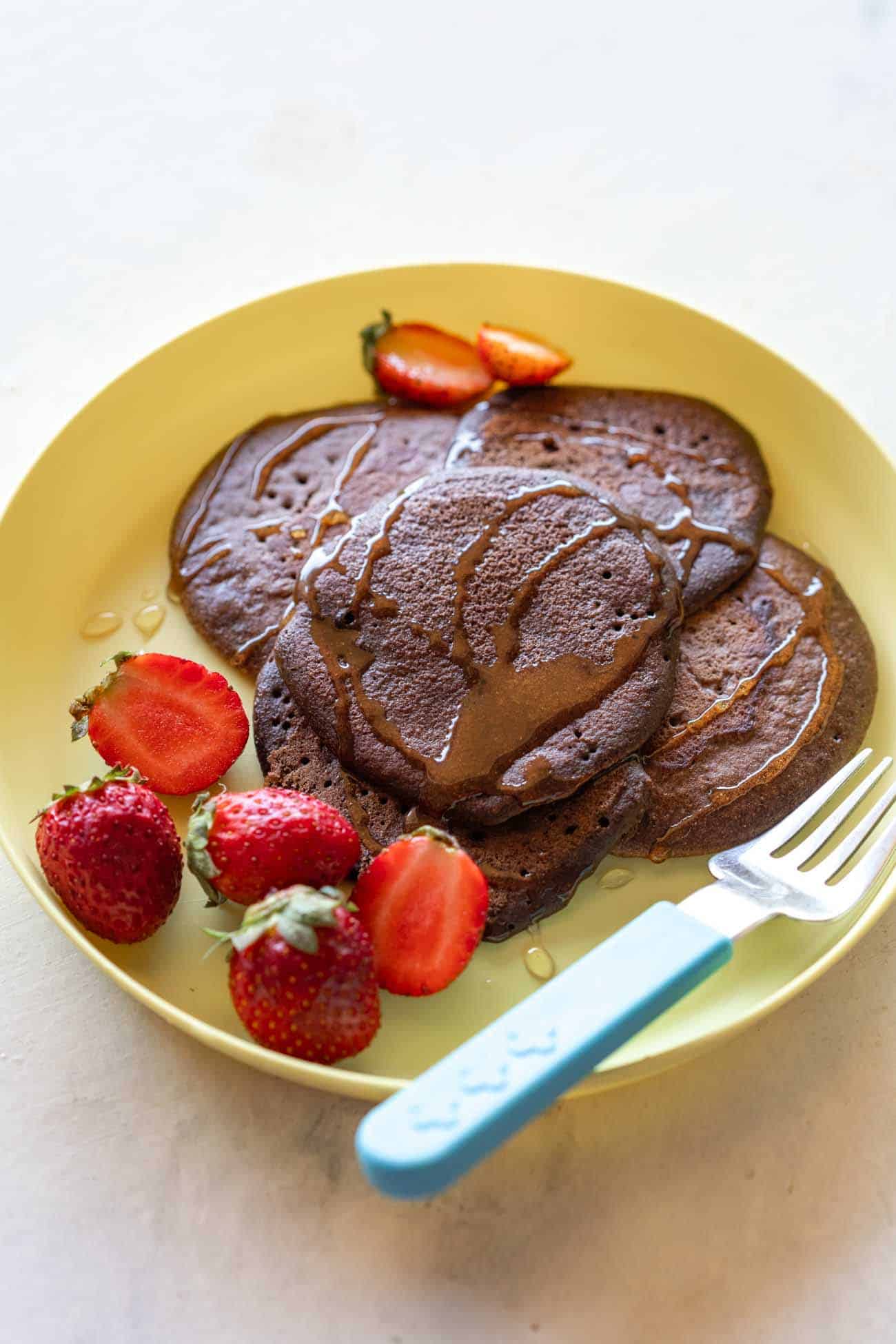 Chocolate Ragi Pancakes served on a yellow plate drizzled with honey and served alongside strawberries