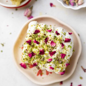 a slice of rasmalai cake garnished with cream, crushed nuts and rose petals on a white plate