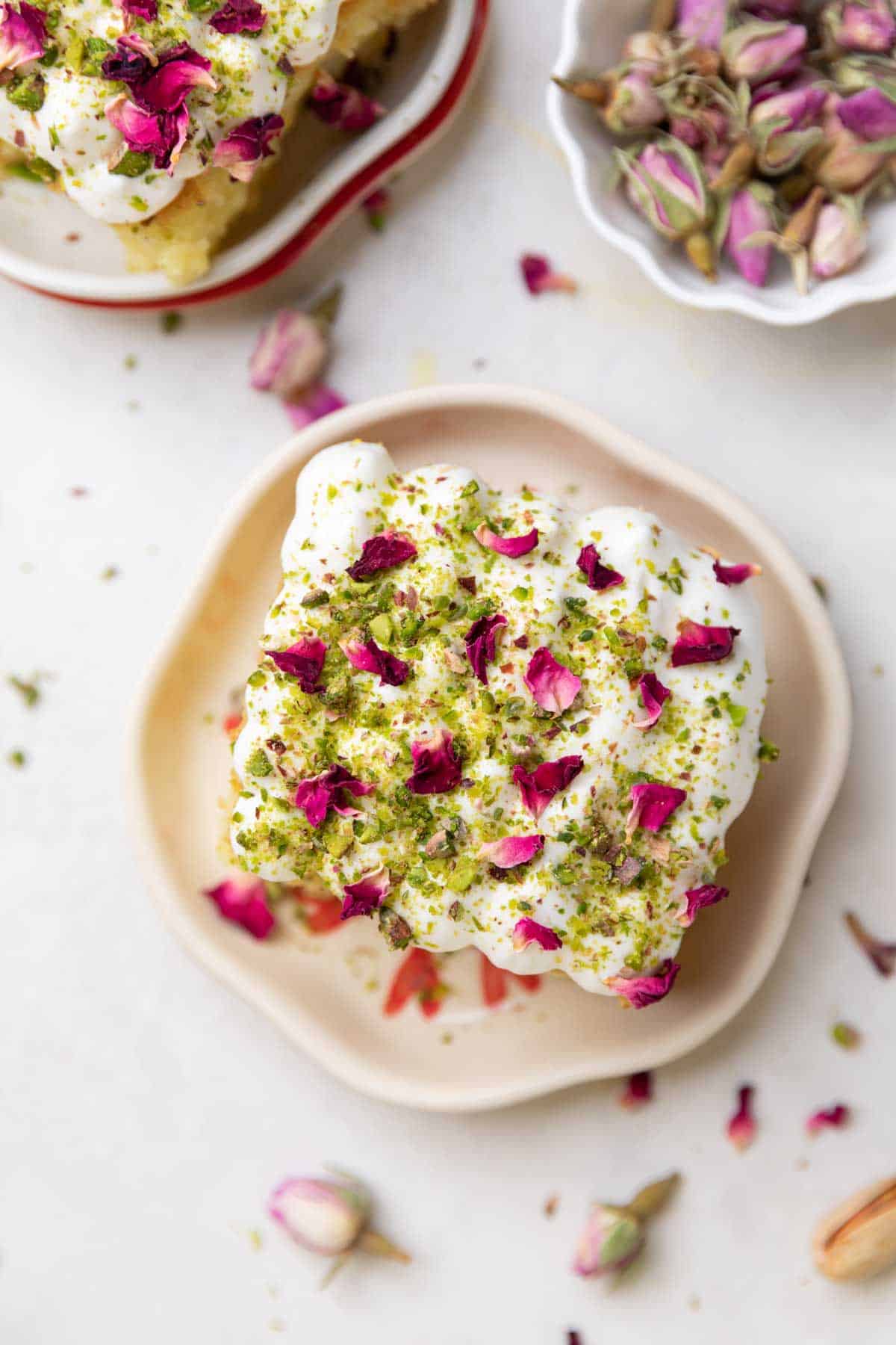 a slice of rasmalai cake garnished with cream, crushed nuts and rose petals on a white plate