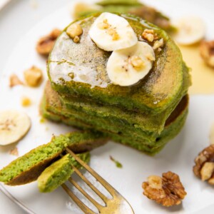 stacked spinach banana pancakes topped with banana slices, walnuts and honey and served with a fork