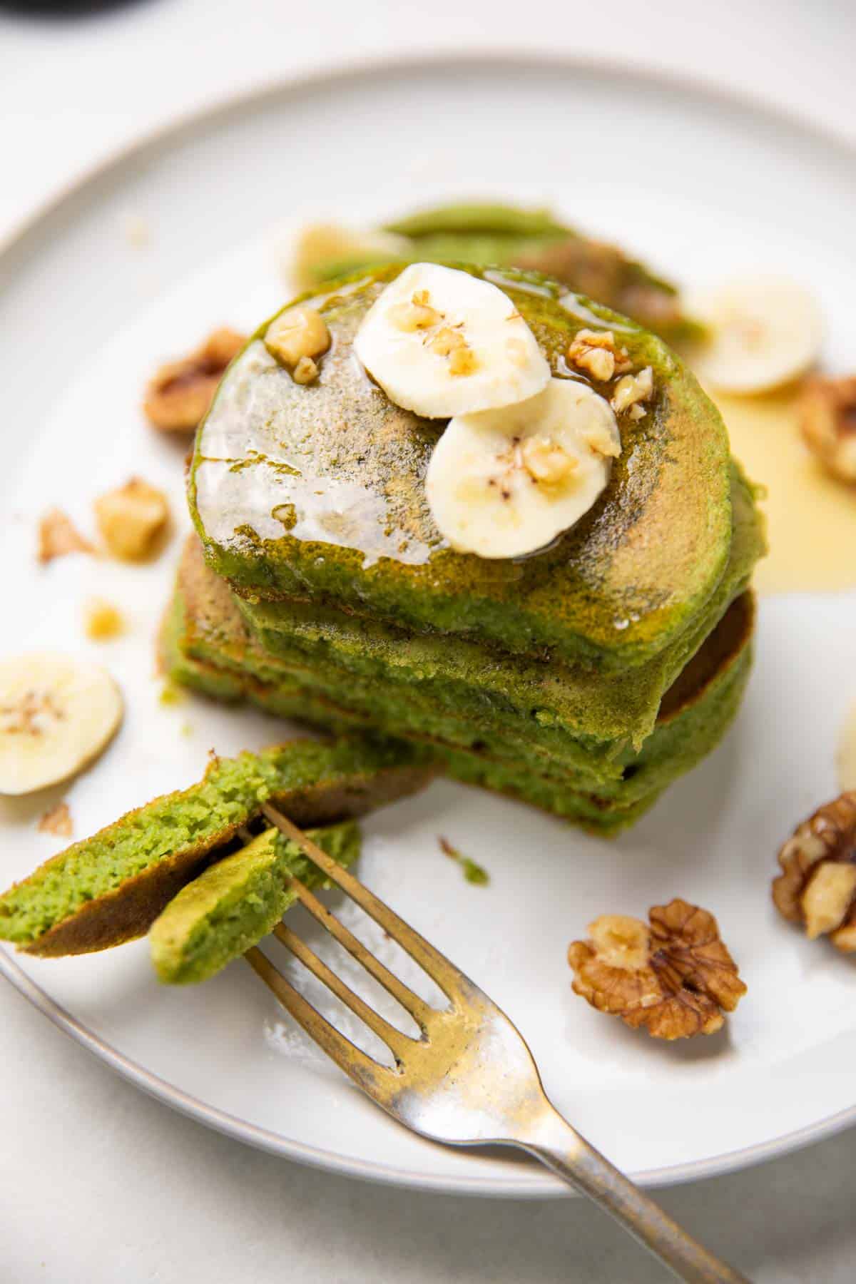stacked spinach banana pancakes topped with banana slices, walnuts and honey and served with a fork